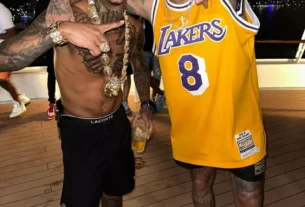 Dois homens posam sorrindo em um ambiente descontraído, possivelmente uma festa em um barco. Um deles está sem camisa, usando bermuda preta, correntes e acessórios dourados. O outro veste uma regata amarela do time Lakers, boné preto e chinelo. Ambos fazem MC Poze do Rodo e Neymar gestos com as mãos e mostram as tatuagens nos braços e pernas.
