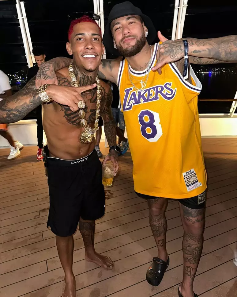 Dois homens posam sorrindo em um ambiente descontraído, possivelmente uma festa em um barco. Um deles está sem camisa, usando bermuda preta, correntes e acessórios dourados. O outro veste uma regata amarela do time Lakers, boné preto e chinelo. Ambos fazem MC Poze do Rodo e Neymar gestos com as mãos e mostram as tatuagens nos braços e pernas.