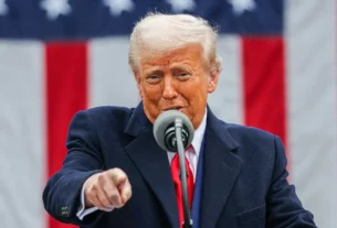 Trump solta o verbo contra o Irã e manda todo mundo vazar de Teerã Homem de cabelos grisalhos, pele alaranjada e expressão séria fala ao microfone, apontando diretamente para a câmera. Ao fundo, há uma grande bandeira dos Estados Unidos com listras vermelhas e brancas e estrelas brancas sobre fundo azul.