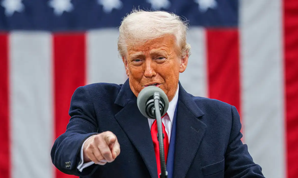 Trump solta o verbo contra o Irã e manda todo mundo vazar de Teerã Homem de cabelos grisalhos, pele alaranjada e expressão séria fala ao microfone, apontando diretamente para a câmera. Ao fundo, há uma grande bandeira dos Estados Unidos com listras vermelhas e brancas e estrelas brancas sobre fundo azul.