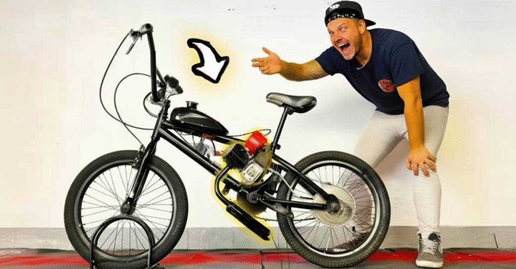 Homem sorridente, vestindo boné, camiseta preta e calça clara, aponta animado para uma bicicleta preta equipada com motor, destacando o tanque de combustível com uma seta desenhada na imagem.