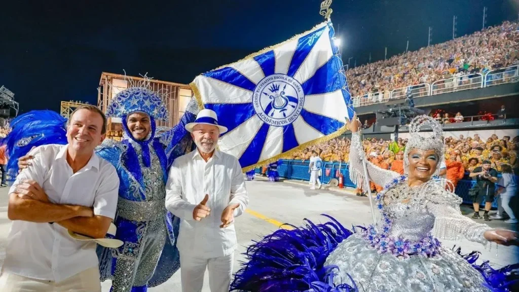 Presidente Lula participando de desfile de carnaval ao lado de integrantes da Escola de Samba Acadêmicos do Tuiuti, segurando a bandeira da agremiação.