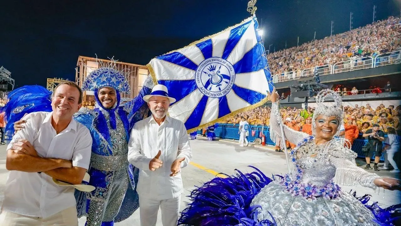 Presidente Lula participando de desfile de carnaval ao lado de integrantes da Escola de Samba Acadêmicos do Tuiuti, segurando a bandeira da agremiação.