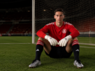 "Goleiro de futebol agachado no campo, usando uniforme vermelho, luvas brancas e chuteiras prateadas, com a rede do gol ao fundo."