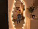 "Espelho de corpo inteiro com iluminação LED em formato orgânico, em um quarto moderno com cama, plantas e decoração minimalista."