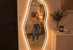 "Espelho de corpo inteiro com iluminação LED em formato orgânico, em um quarto moderno com cama, plantas e decoração minimalista."